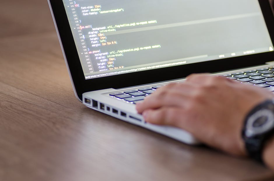 The image shows a silver laptop open on a light-colored wooden desk, displaying lines of code on its screen, indicative of programming or software development work. Next to the laptop, there is an ope